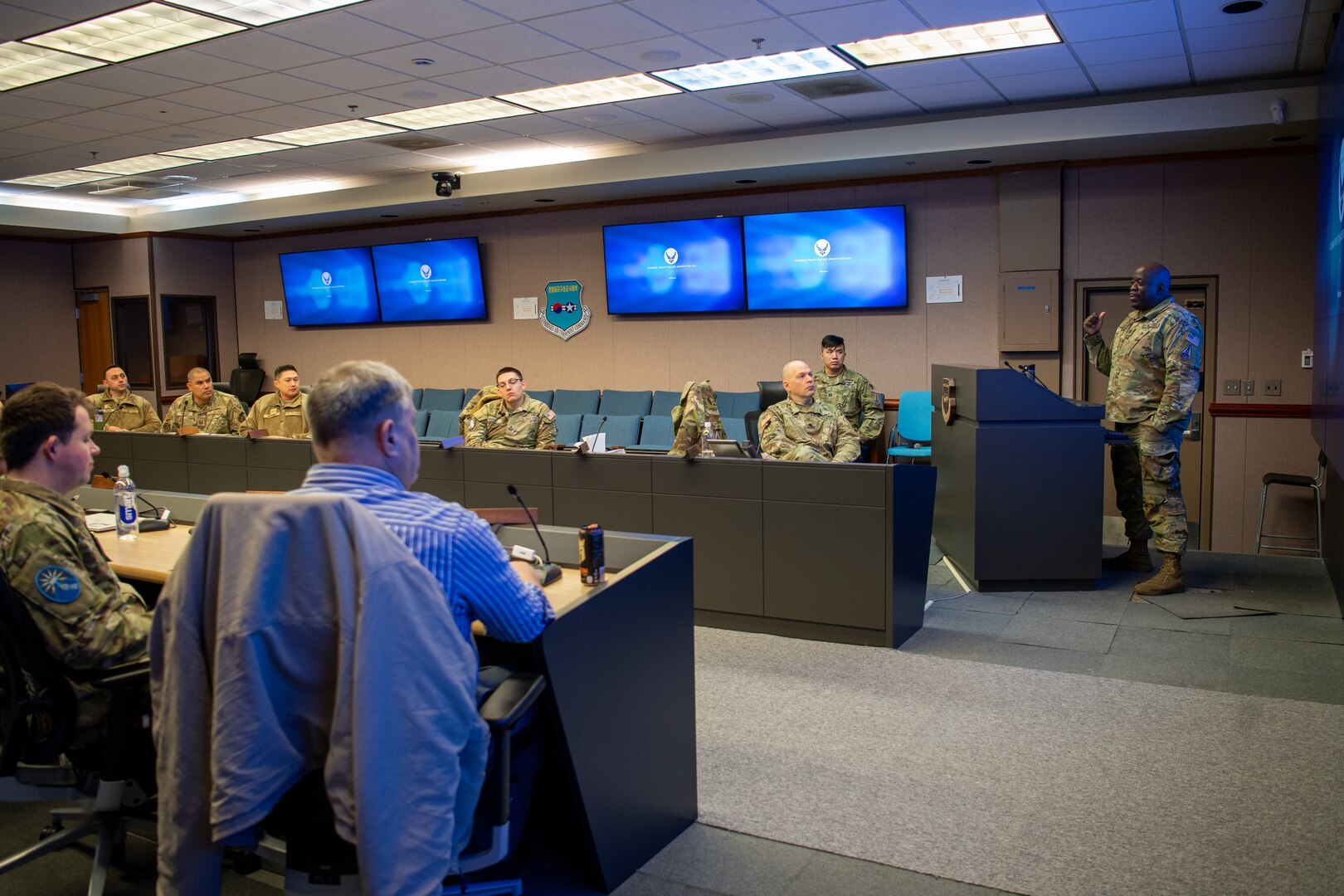 Eight U.S. Space Force Guardians and civilians sit around a conference room table, while one Guardian in uniform stands behind a podium on the right side of the room, and several screens on the wall project slides the audience.