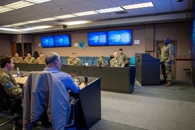 Eight U.S. Space Force Guardians and civilians sit around a conference room table, while one Guardian in uniform stands behind a podium on the right side of the room, and several screens on the wall project slides the audience.