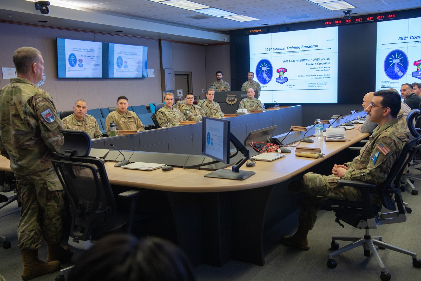A dozen Space Force members sit in around a conference table, being addressed by one man on the left and head of the table, with a large screen in the background showing slides.