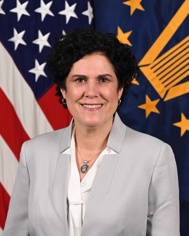 A woman in business attire poses in front of two flags.