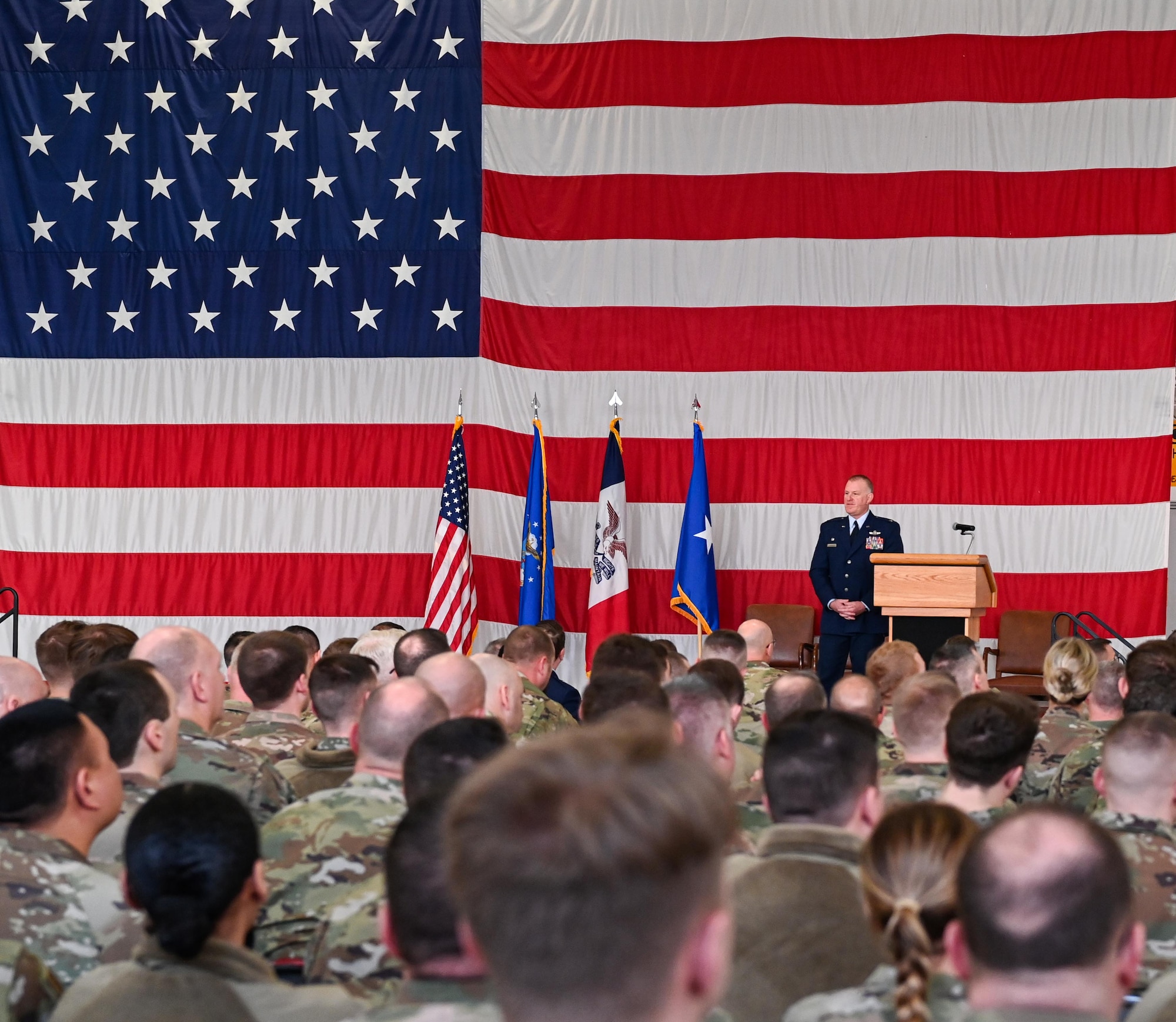 Crowd of airmen watch as Air Force colonel in blue uniform gives a speech on a stage in front of giant U.S. flag.