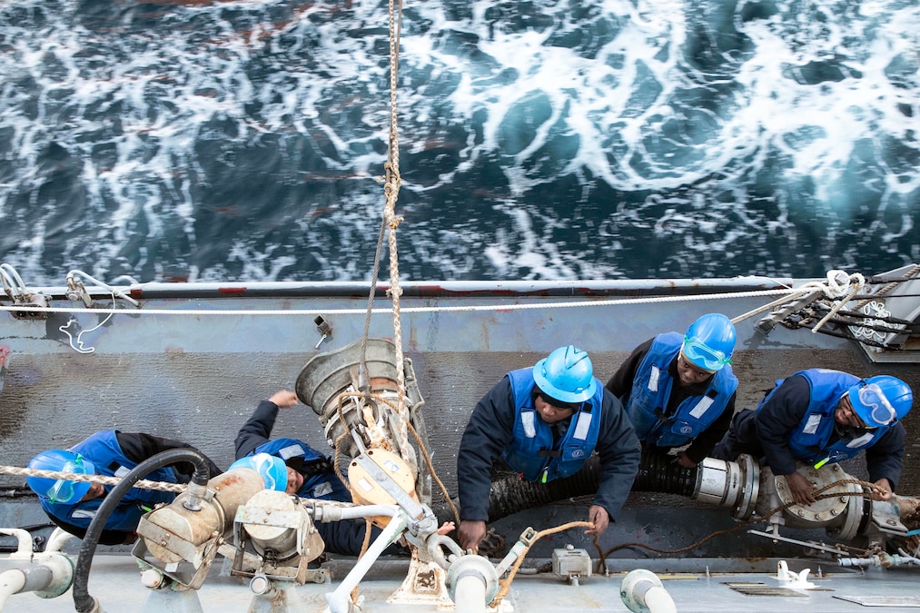 Five sailors wearing dark blue vests and light blue hardhats work on the side of a ship at sea as seen from above.