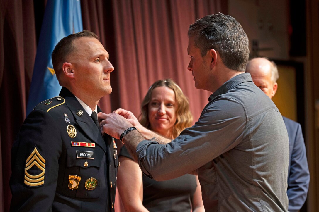 A person pins a medal on a soldier in uniform in front of a red curtain while two people watch from behind.