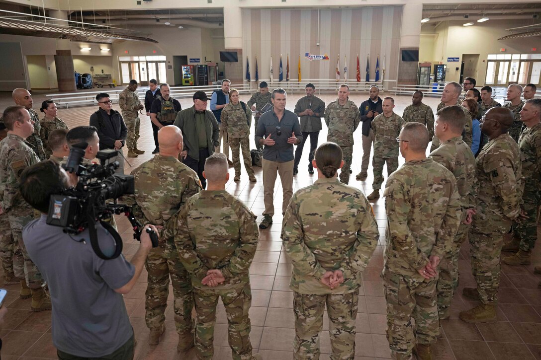 A large group of soldiers stand in a circle around a person speaking in a spacious, tan room.