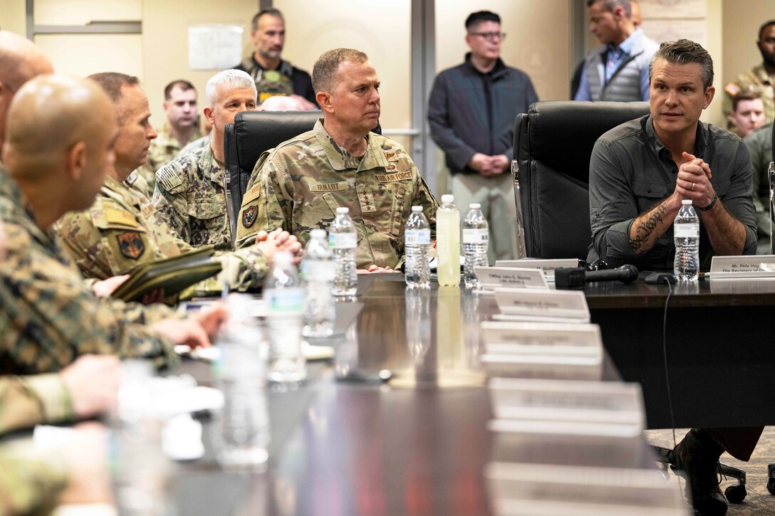 A group of soldiers sits around a table in a bright room while one person clasps their hands together.