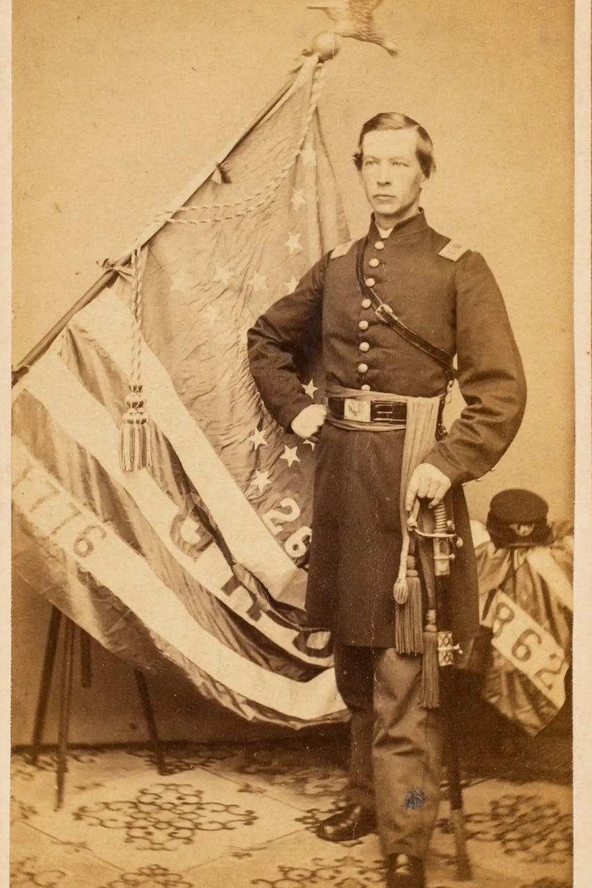A man in a Civil War-era military uniform stands in front of an American flag.