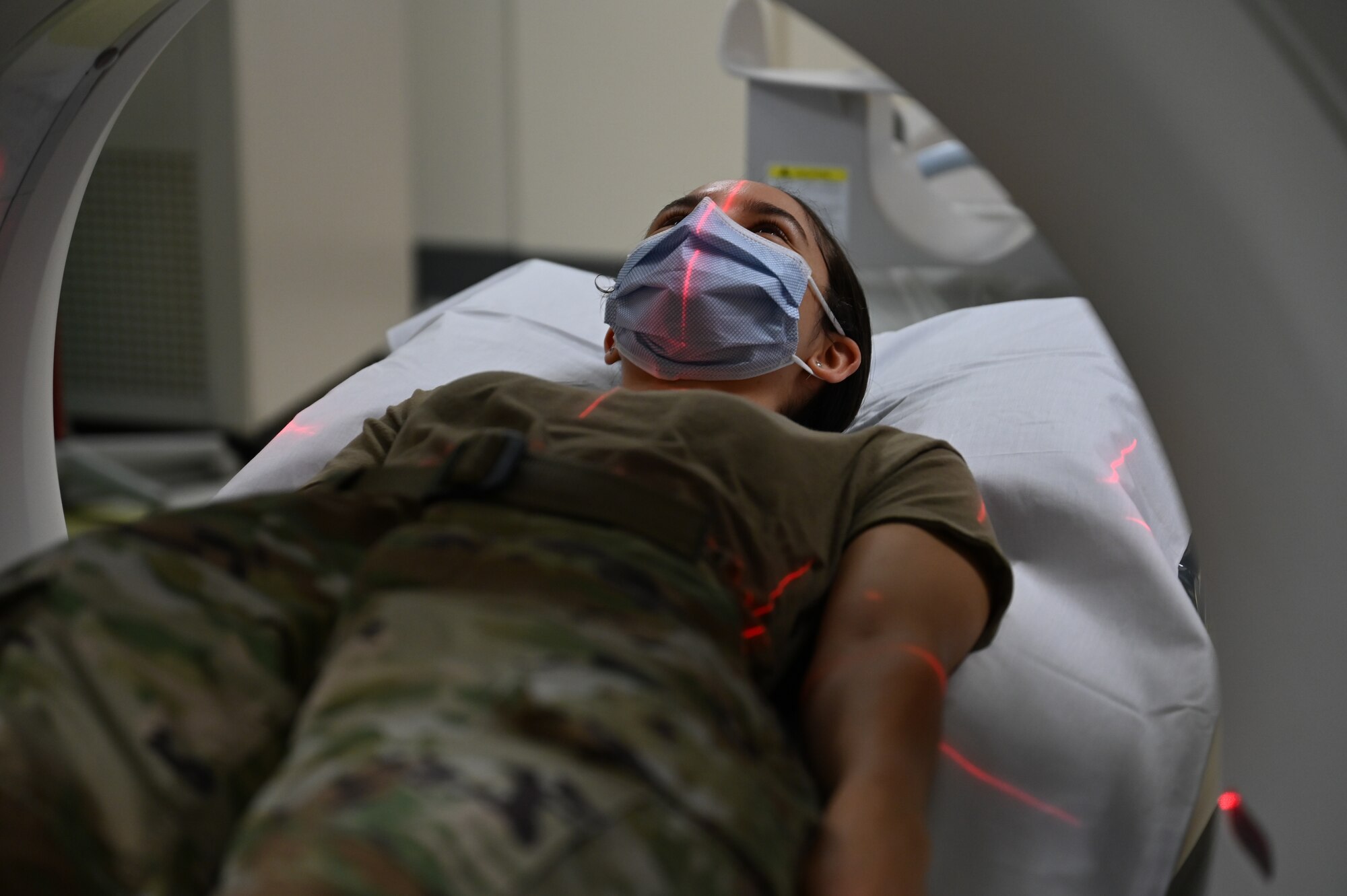 Photo of a uniformed woman in a CT machine with red lines across her face and chest. She is wearing a mask.