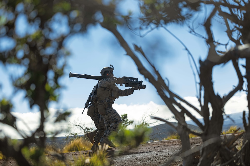 Two Marines, seen through branches against a blue sky, run together on gravel terrain with a large weapon.