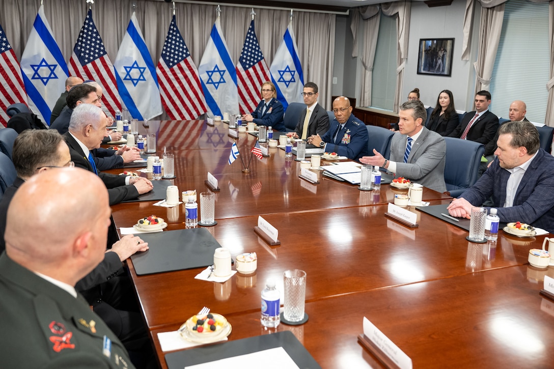 A group of people sit around a large conference room table with the U.S. and Israeli flags in the background.