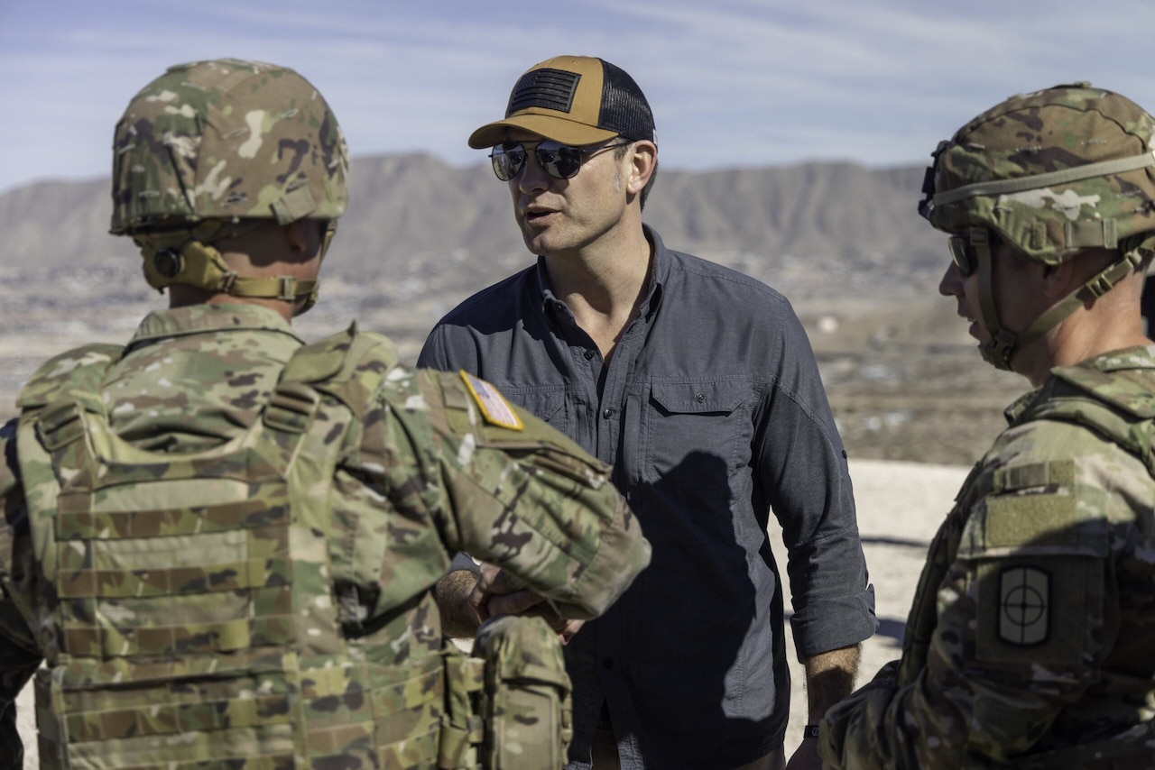 A man in civilian attire, sunglasses and a baseball hat is talking to two people in camouflage uniforms.