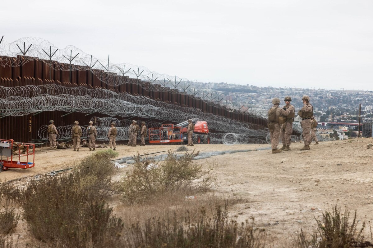 Service members in camouflage uniforms are standing near a large outdoor wall that is reinforced with barbed wire.