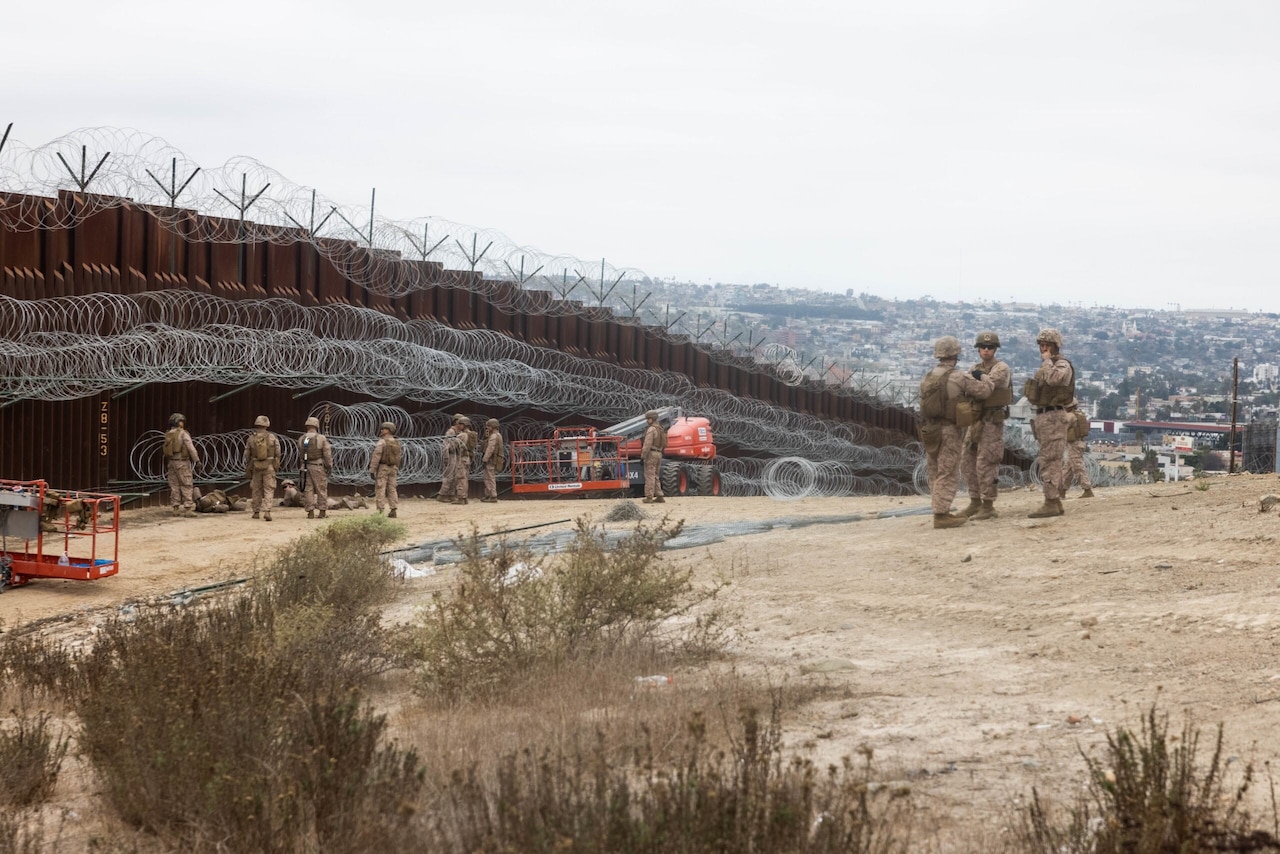 Service members in camouflage uniforms are standing near a large outdoor wall that is reinforced with barbed wire.