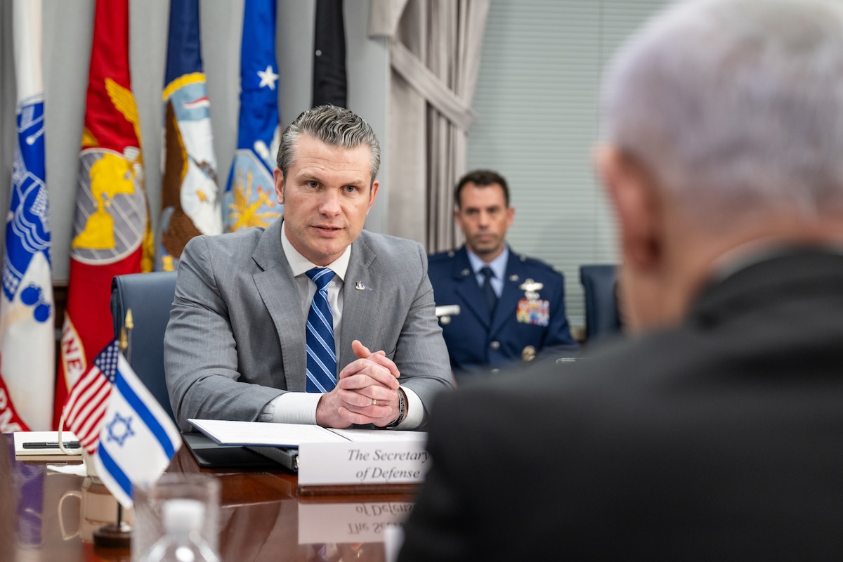 Defense Secretary Pete Hegseth sits at a table across from another person while a third person sits behind him with flags in the background.