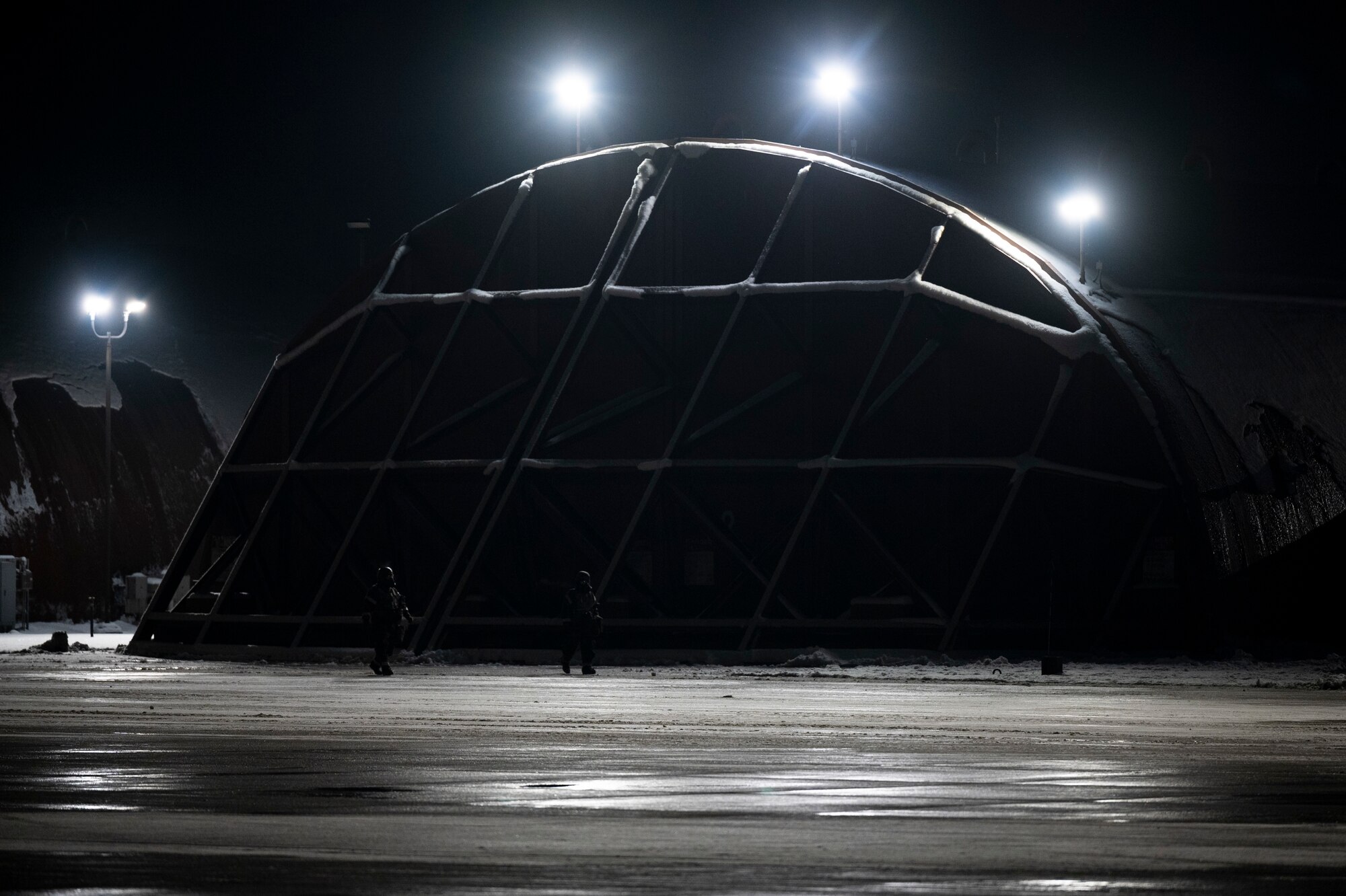 Two Airmen in protective gear look for signs of contamination while outside at night with a hangar in the background.