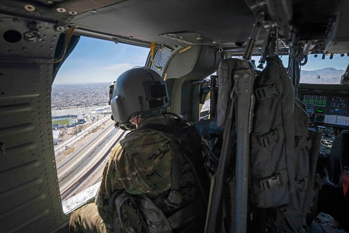 A pilot looks out of a helicopter at a highway under blue skies.