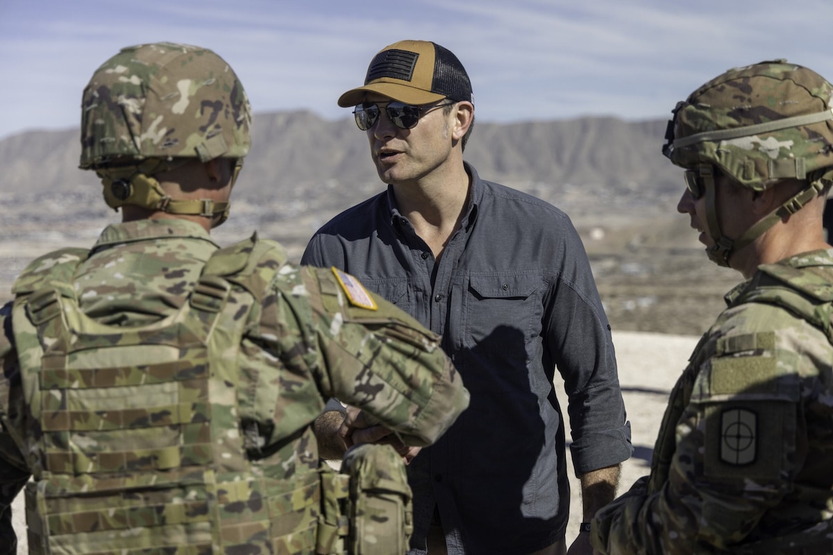 A man in civilian attire, sunglasses and a baseball hat is talking to two people in camouflage uniforms.