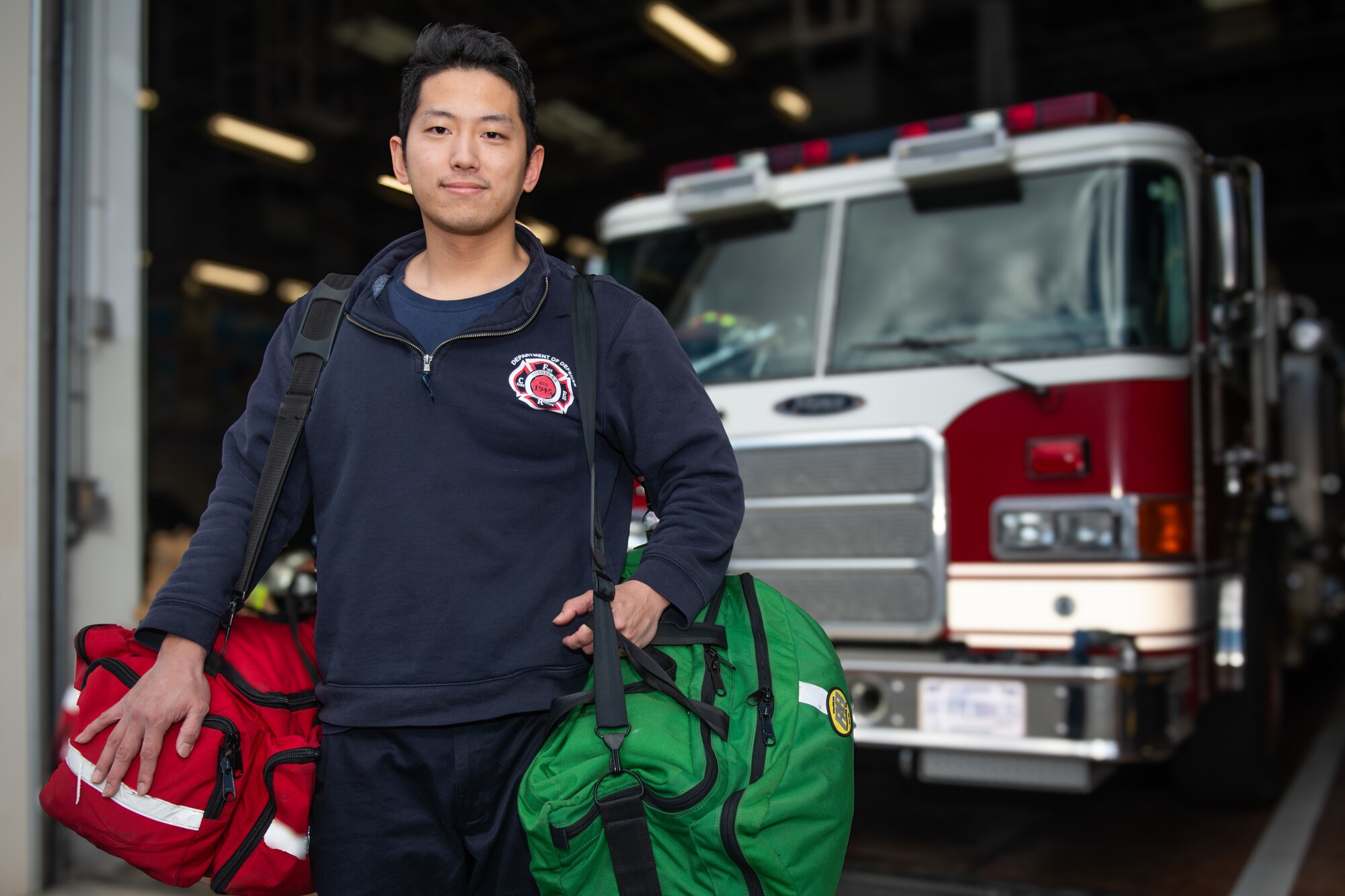 A374th Civil Engineer Squadron firefighter holds red and green medical supply bags in front of a red fire truck.