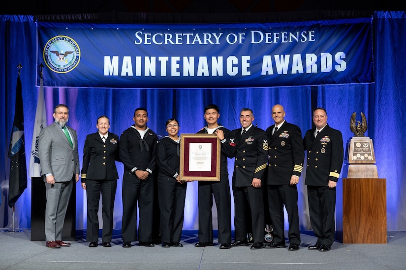 A man and woman in uniform in the middle hold up a framed letter next to six others on stage. A banner above them reads “Secretary of Defense Maintenance Awards.”