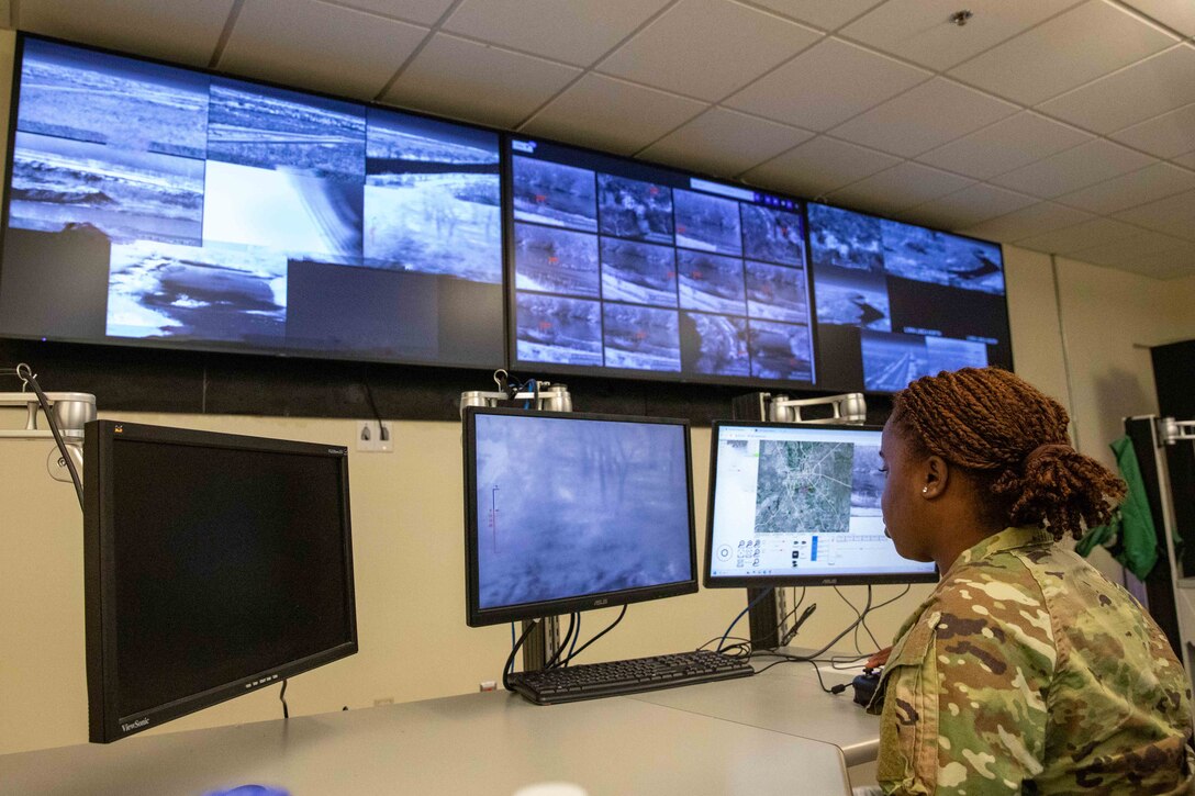 A soldier looks at six computer monitors while seated at a desk inside a room.