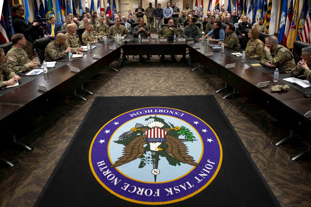 Military officials and soldiers sit around a large table with a carpet in the middle of the United States seal.