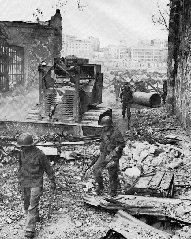 Troops walk along the street of a ruined urban area.