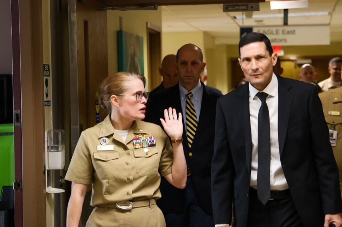 A female in uniform leads two men in suits down a hospital hallway.