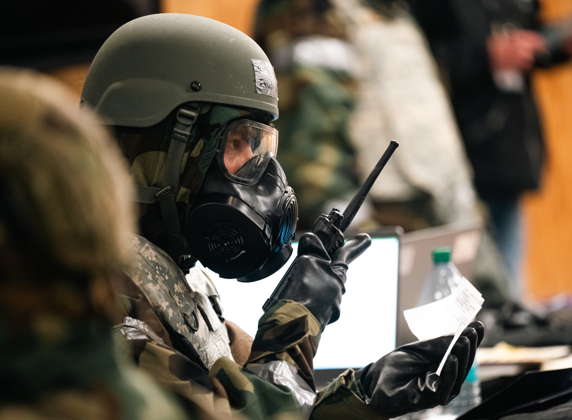 A man in cbrn gear holds a radio while sitting in a room