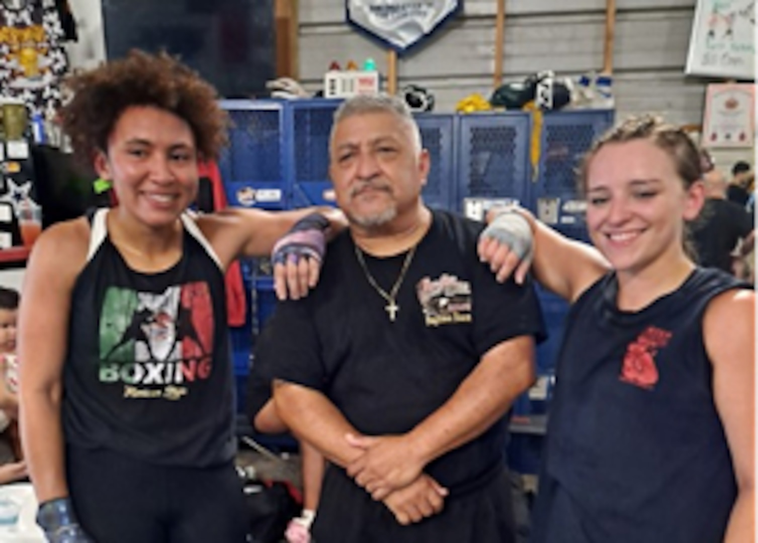 A man stands with two young female boxers, one on either side of him. the background is the inside of a boxing gym.
