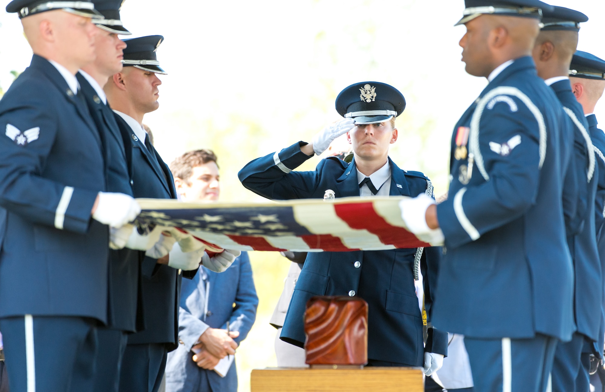 several members of the Air Force Honor Guard uniform ready to begin folding the US flag during a funeral service, while one guard salutes.