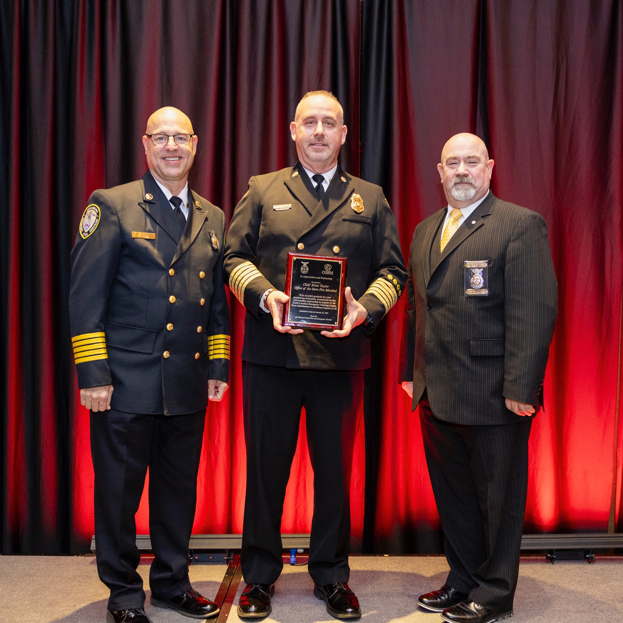 Chief Daryl D. Cook, 145th Airlift Wing installation fire chief, left, and Charles B. Loveday, Air National Guard command fire chief, right, present Brian Taylor, North Carolina State Fire Marshal, middle, with a plaque in recognition of a longstanding partnership with the 145th Airlift Wing at the Mid-Winter Fire Chiefs Conference banquet at the Embassy Suites in Concord, N.C., Jan. 31, 2025.