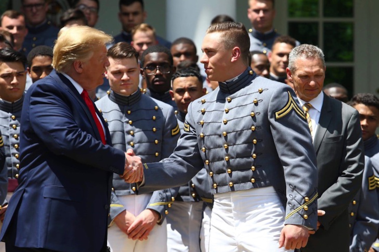 A man in a suit shakes hands with a military cadet in a dress uniform.