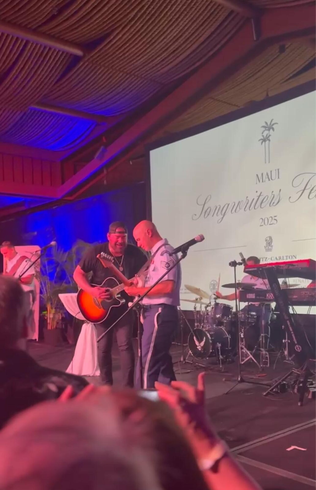 A uniformed soldier plays guitar with a civilian guitarist on a hotel stage.