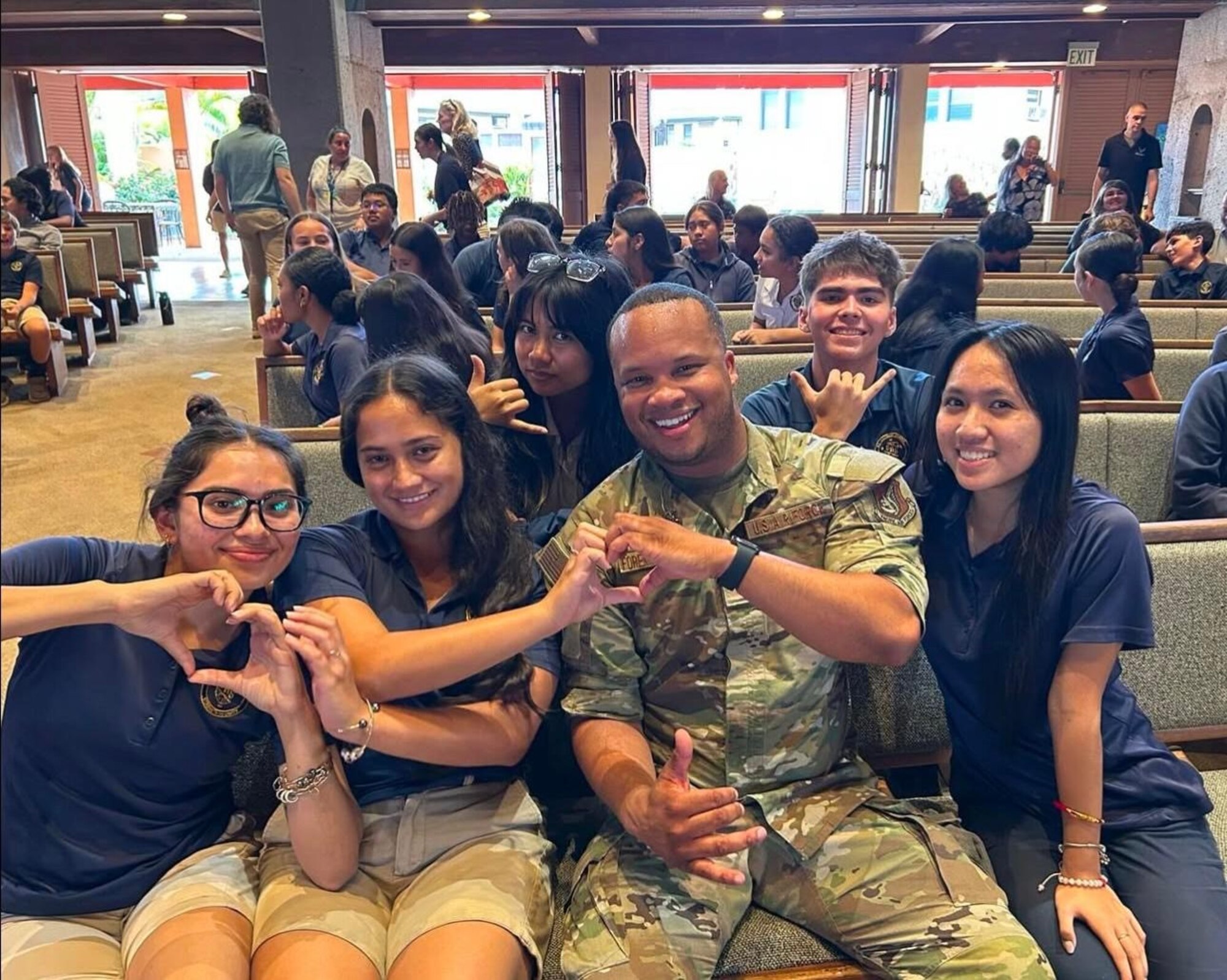 A uniformed soldier makes a heart sign with several students surrounding him while seated in a school auditorium.