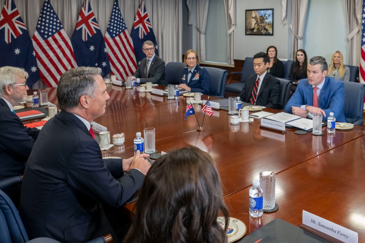 Multiple individuals sit across a table from one another. U.S. and Australian flags are posted in the rear.