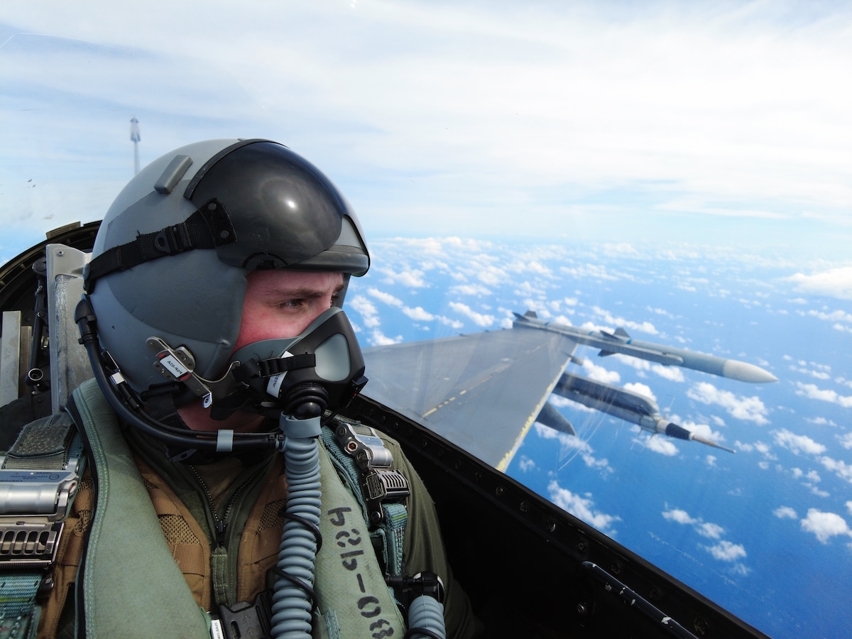 An Airman sits in the back seat of a fighter jet and looks out across the ocean, preparing to document the flight.