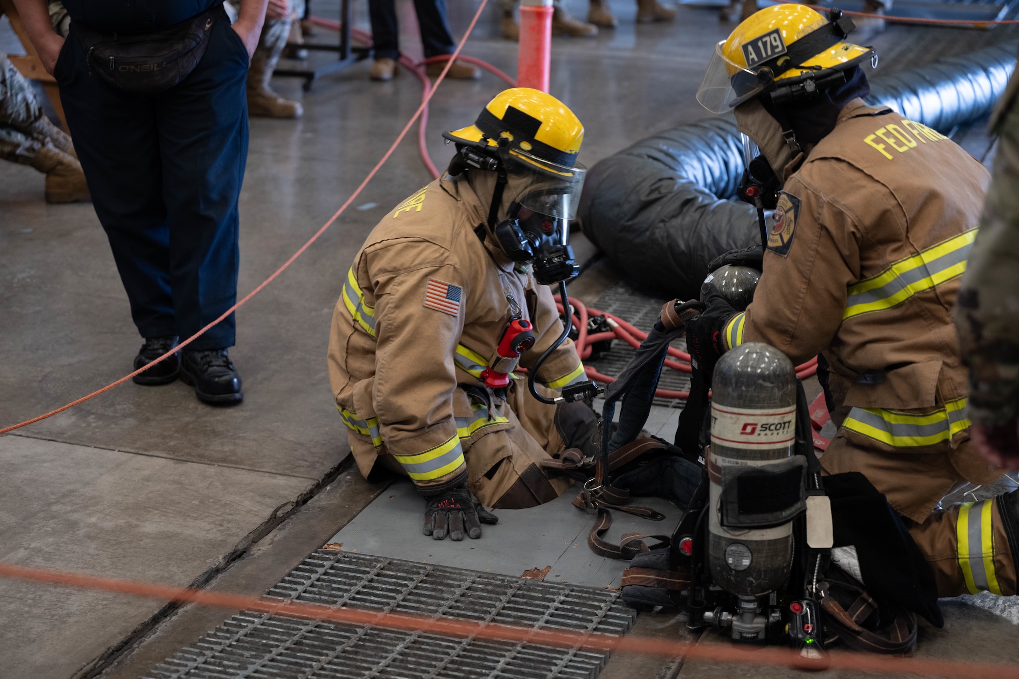 A firefighter attempts to go down a small hole. With another firefighter kneeling next to him.