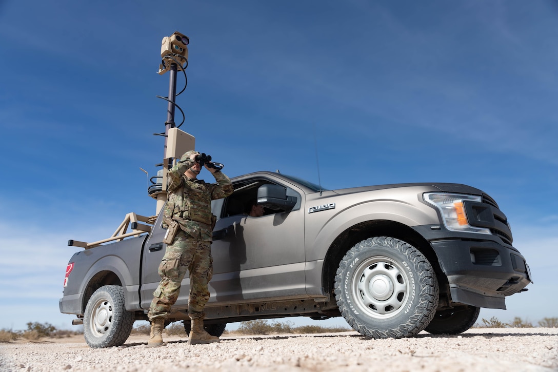 A soldier standing next to a vehicle with a communications tower, looks through binoculars.