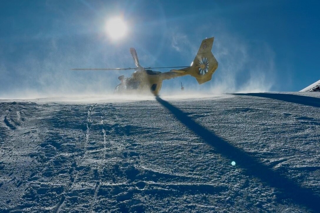 A recue helicopter takes off from the top of a snowy hill during a sunny day.