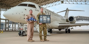 U.S. Air Force Lt. Col. Randell McCart, the 430th Expeditionary Electronic Combat Squadron commander, and Maj. Eric Goodman, a 430th EECS E-11A pilot, hold up a memorial dedicated to two fallen E-11 A pilots in the U.S. Central Command area of responsibility, Jan. 27, 2025, marking the 5th anniversary of an E-11A crash that claimed the lives of two 430th EECS pilots, Lt. Col. Paul “Tabs” Voss and Capt. Ryan “Fogg” Phaneuff.