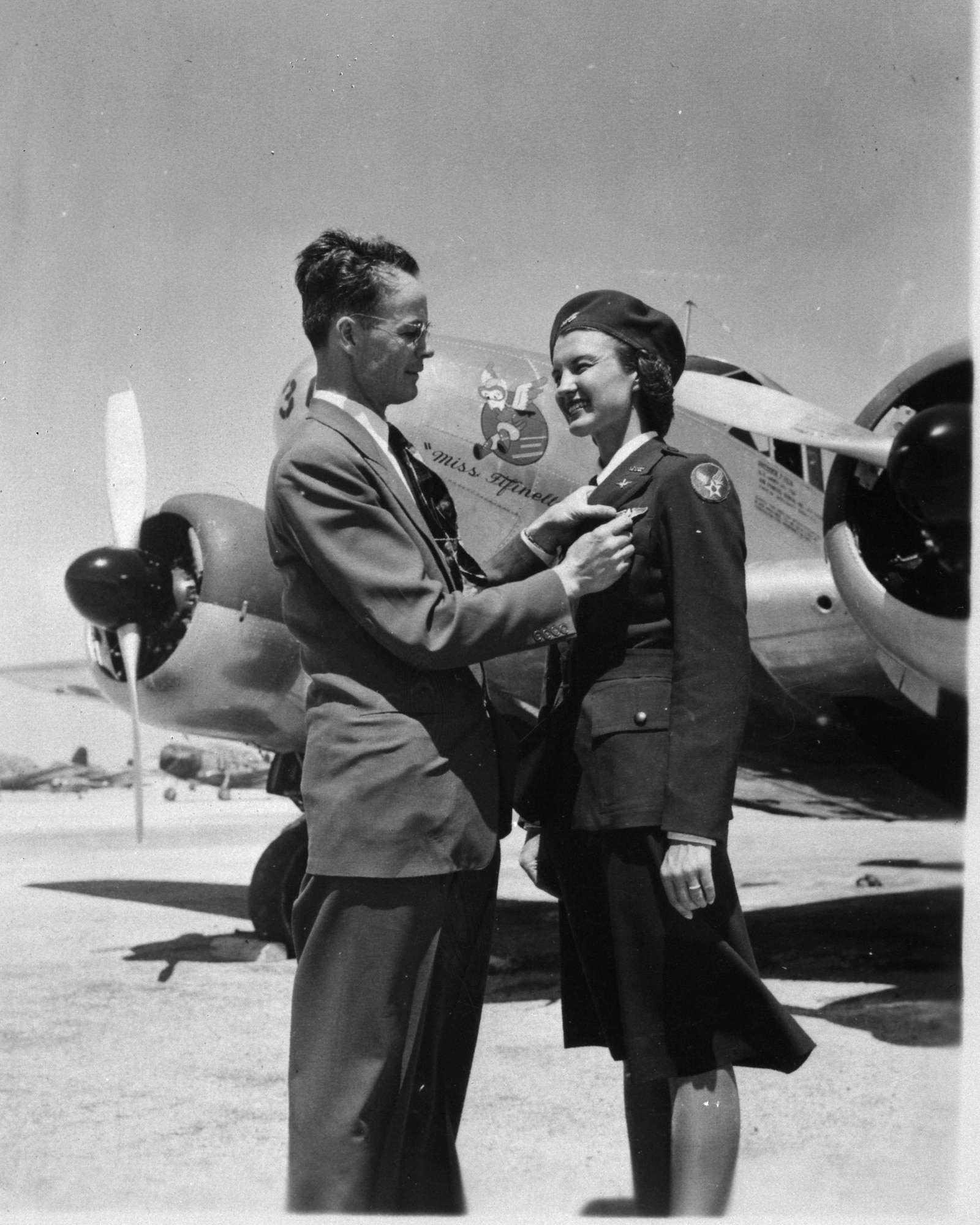 A WASP graduate in uniform standing in front of an aircraft while a man standing next to her is admiring her flight wings attached to her lapel.