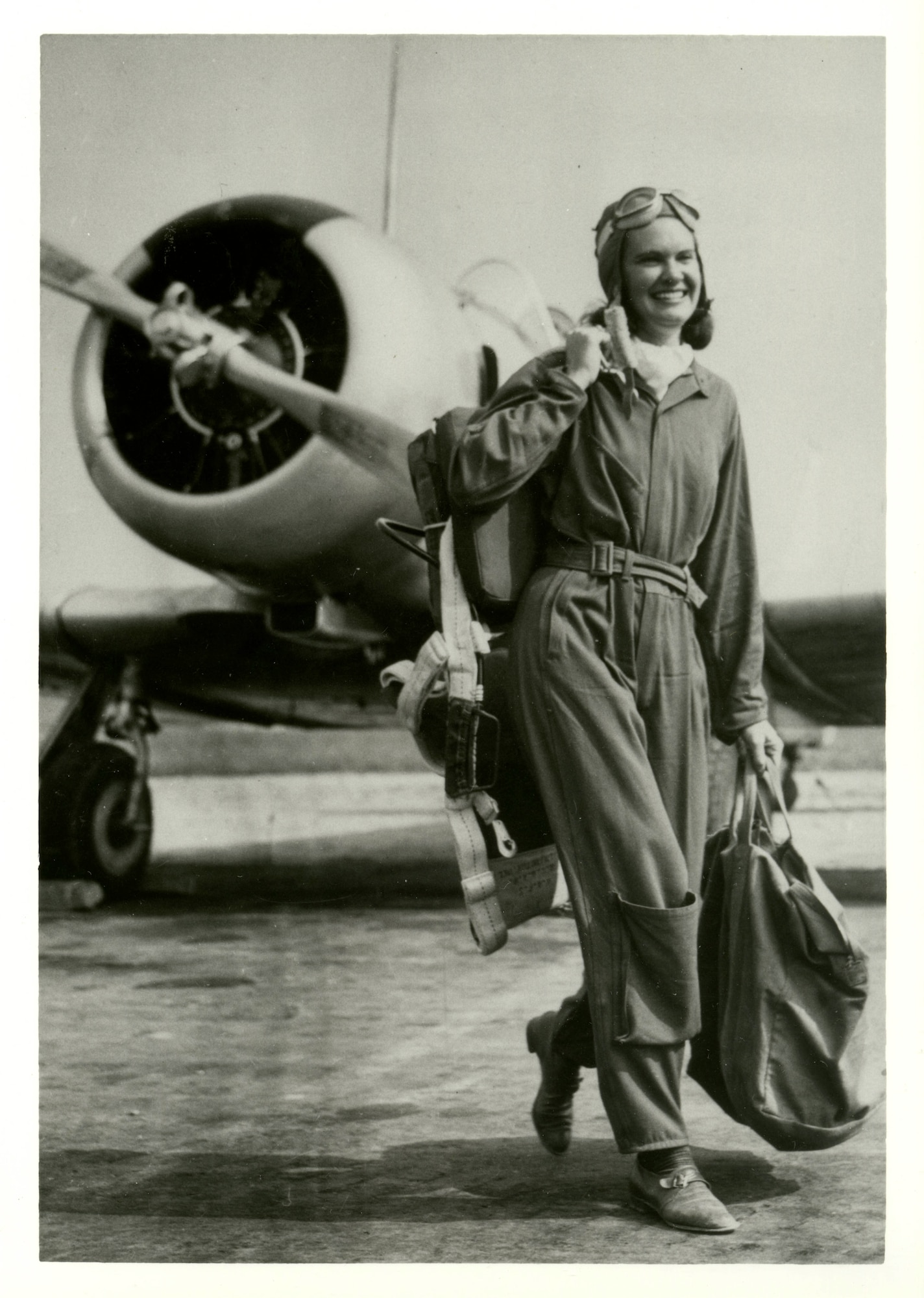 Woman in flight uniform wearing flight cap and goggles on top of her hear, carrying parachute over her shoulder with one hand and duffle bag in other hand as she walks away from aircraft.