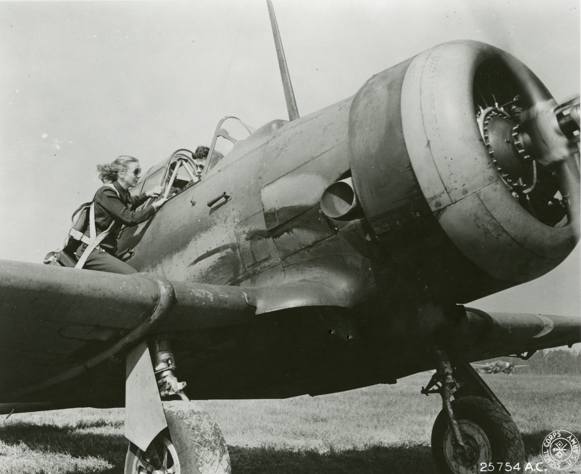 A running airplane with a WASP in the pilot seat speaks to a second WASP standing on the wing.