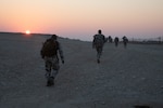 soldiers carrying ruck sacks walk down a dirt road into a sunrise