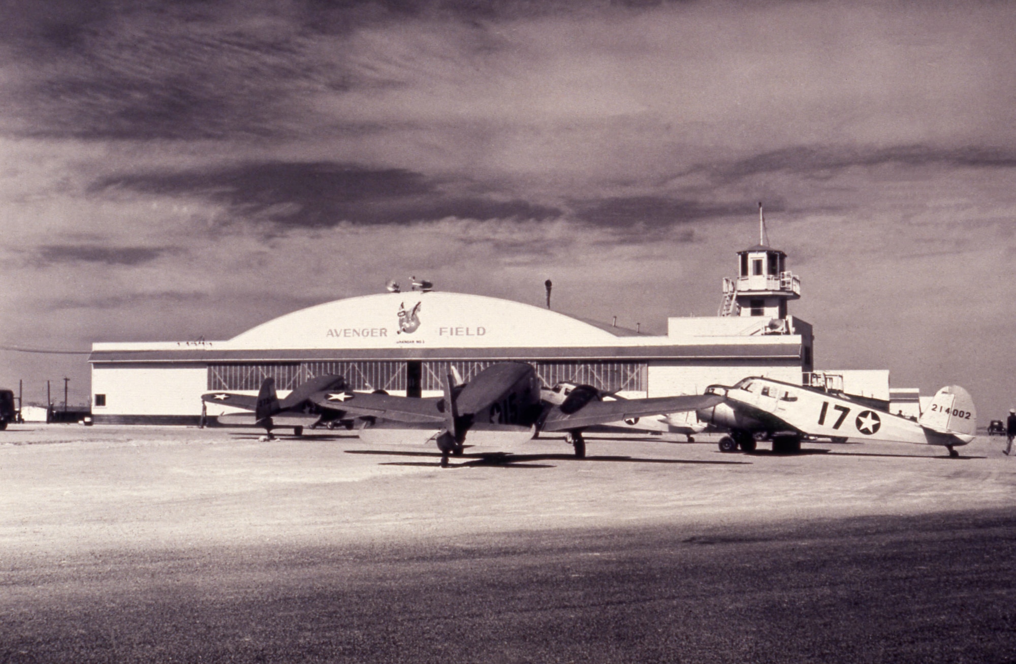 several aircraft sitting outside a hangar with the words Avenger Field displayed with the graphic of Fifinella, the official mascot of the Women Airforce Service Pilots.