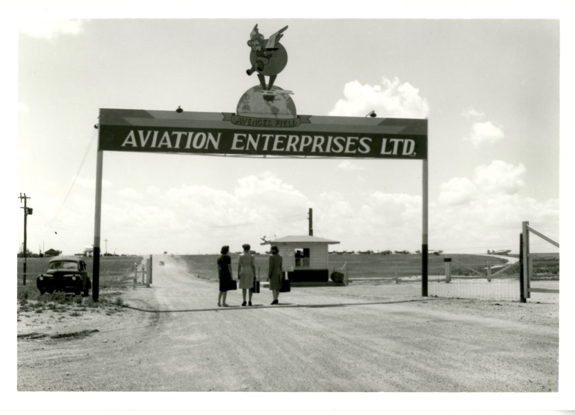 Three women in dresses carrying suitcases stand in the road under a sign that says Aviation Enterprises Ltd.