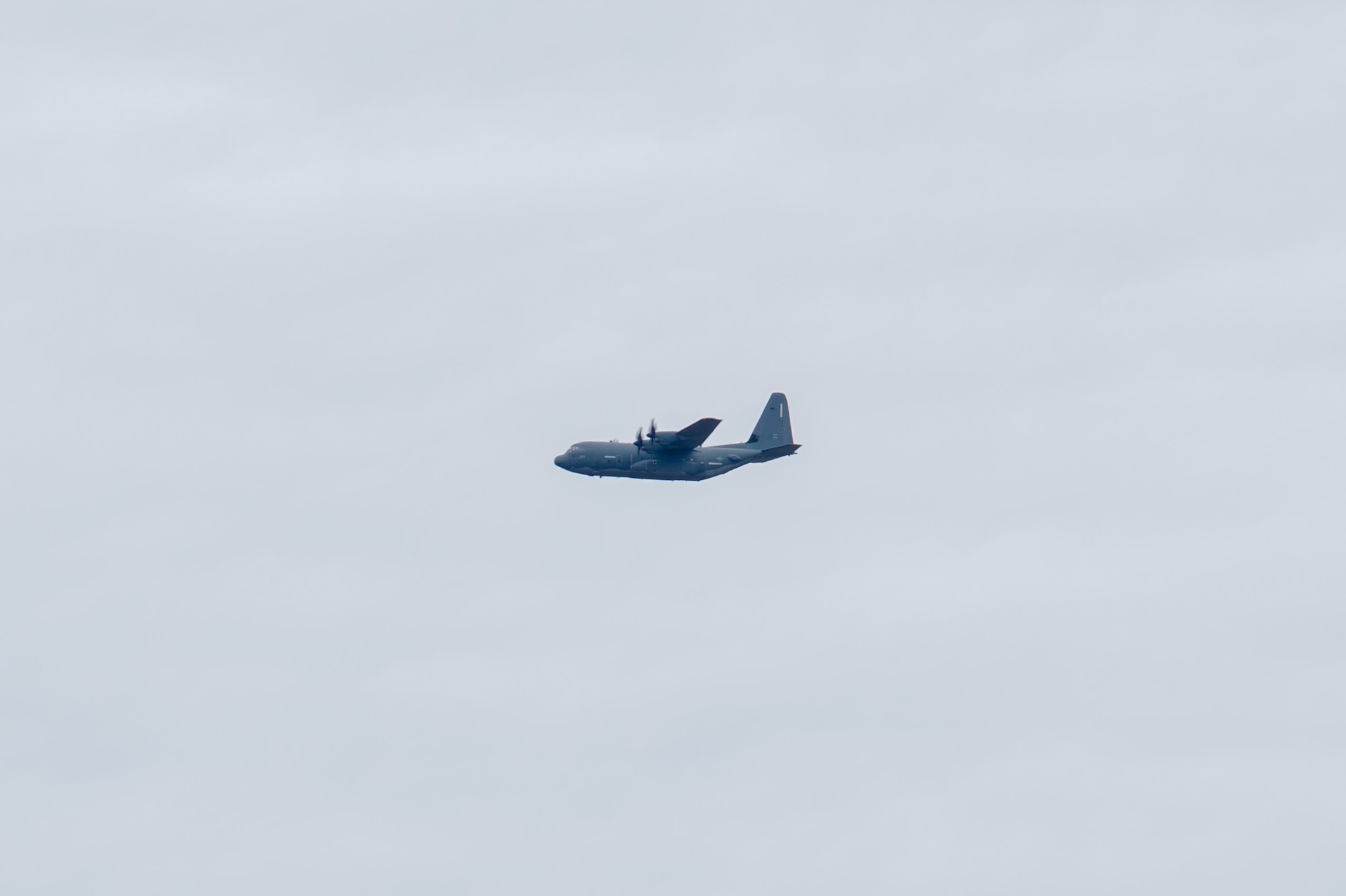 A J-model C-130 aircraft is flying in the distance on a cloudy day.