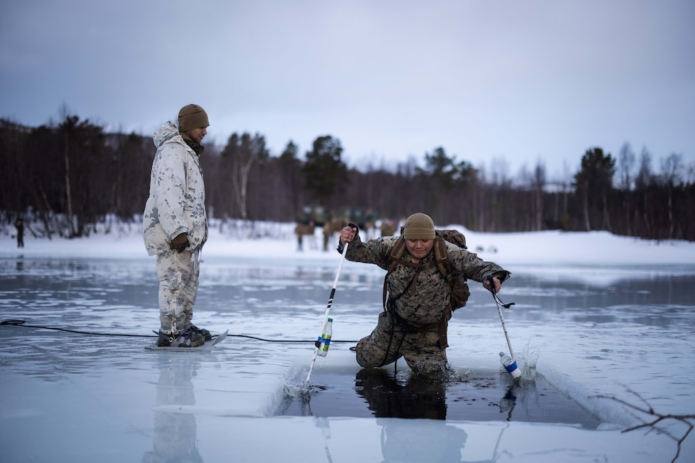 A Marine using ski poles with attached empty water bottles jumps into a square of water in an icy lake while another Marine watches.