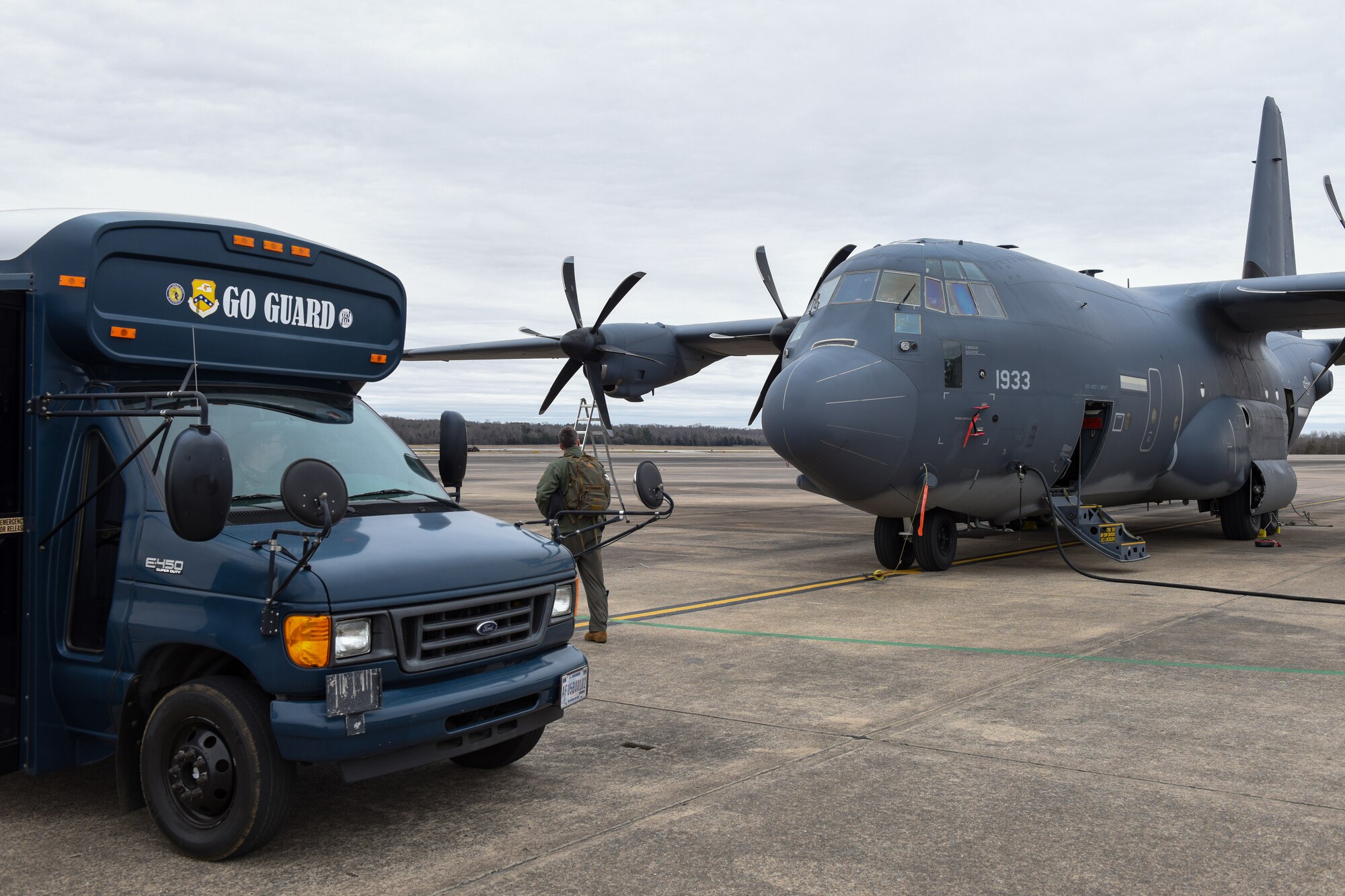 A bus with the189th Airlift Wing logo and the phrase "GO GUARD" on its front is parked in front of a J-model C-130 going through routine inspection on a flightline