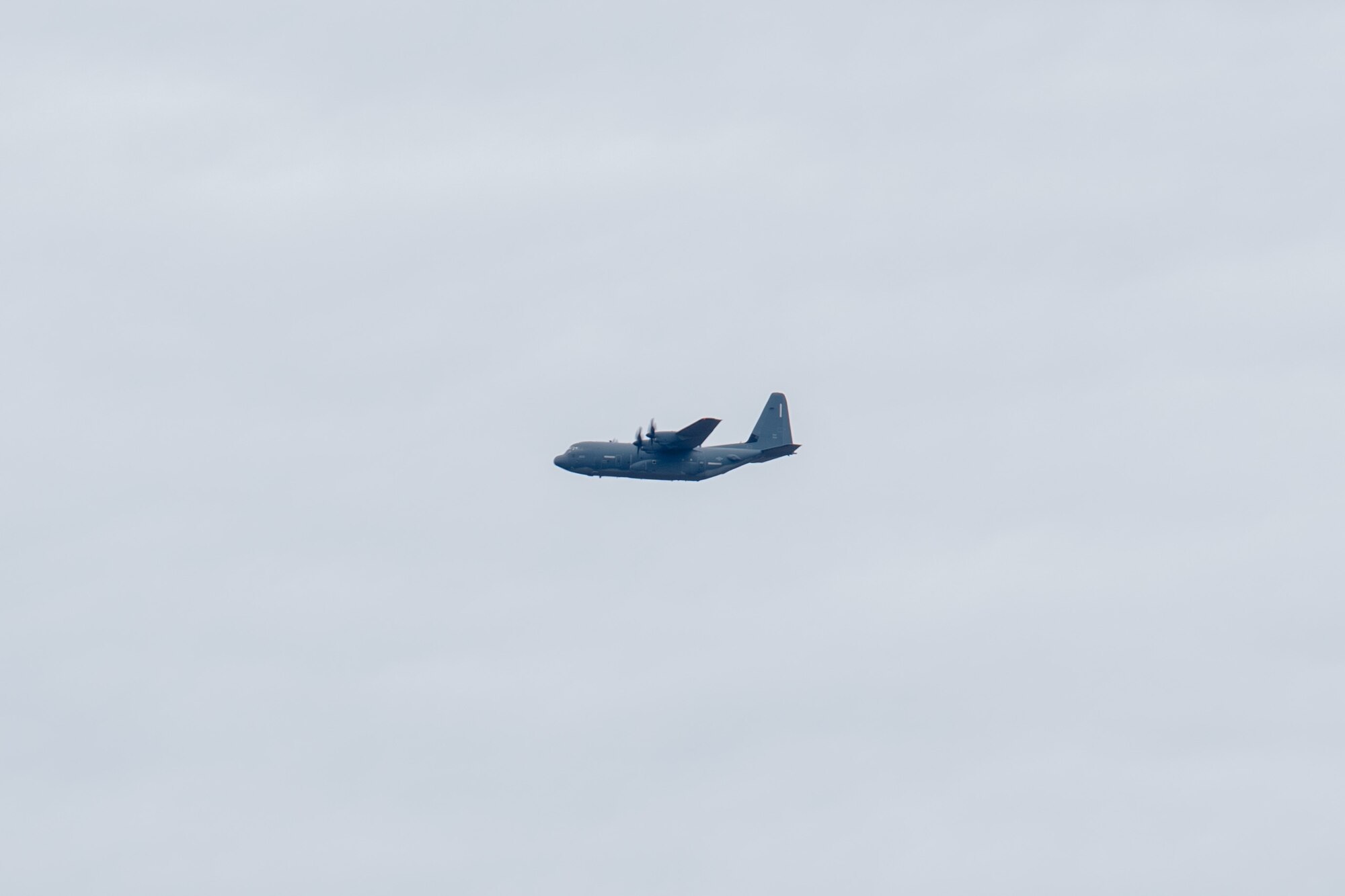 A J-model C-130 aircraft is flying in the distance on a cloudy day.