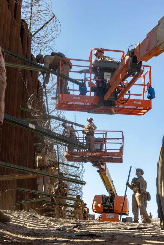 Marines on orange lifts work on barbed wire attached to a fence as fellow Marines work below.